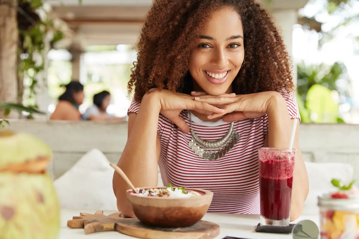 Portrait of happy dark skinned young female with frizzy hair, eats something and drinks smoothie, spends free time with boyfriend or friend, enjoys summer vacations in tropical country on island